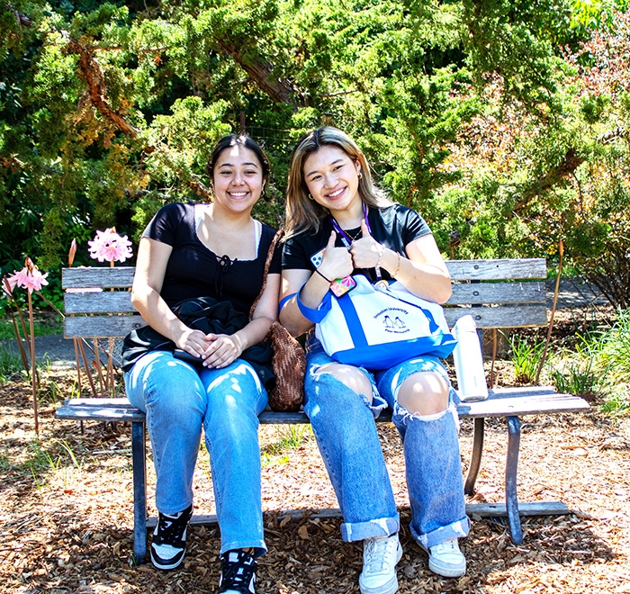 Two young women sitting on a bench and smiling at the camera. One of them giving a two thumbs up gesture of approval..