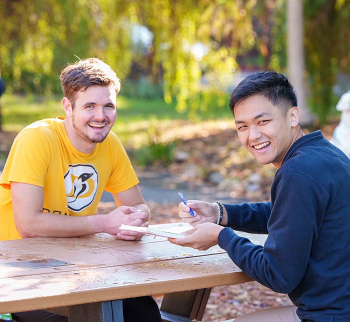 two male students sitting at a picnic table smiling